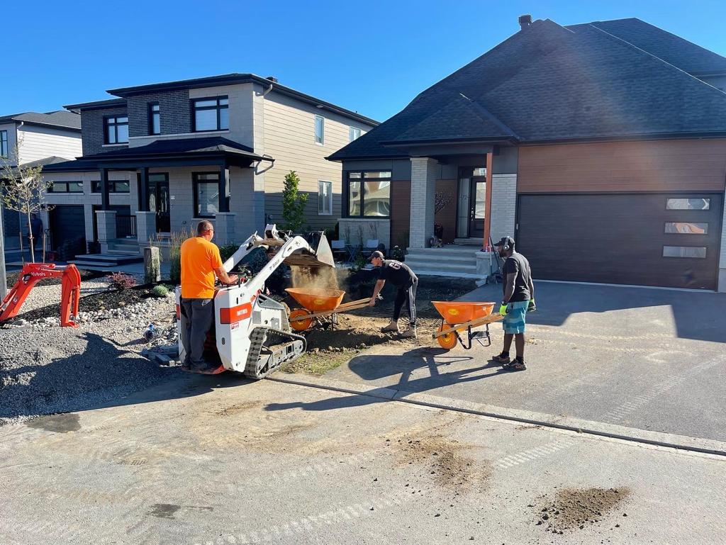 A landscaping team is hard at work in front of a modern home, with one member operating a compact white skid steer loader to move gravel, while others transport materials with wheelbarrows and conduct ground leveling, showcasing a busy and efficient outdoor project site.
