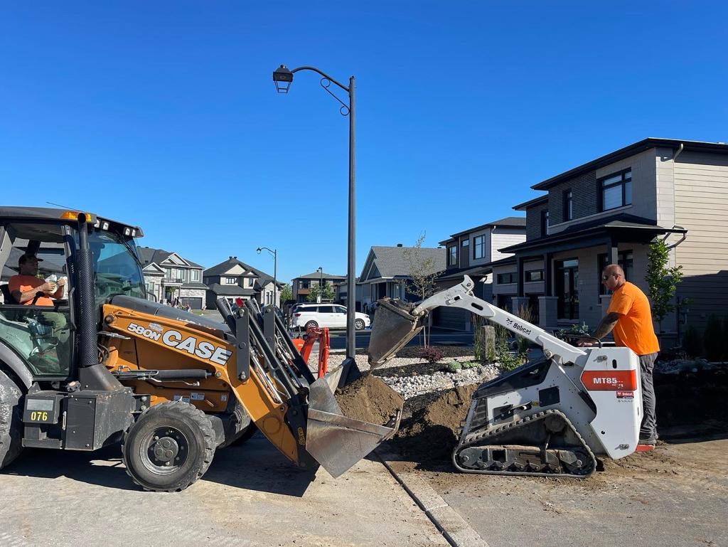 A skilled landscaping crew using heavy machinery on a residential site. An operator is inside a CASE skid steer loader while another worker uses a white Bobcat track loader to move dirt, demonstrating teamwork and machine efficiency in a suburban neighborhood.