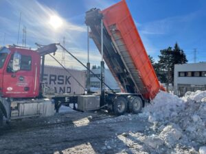 A red dump truck tilts its container upwards, unloading a heap of snow beside a storage container and office buildings.