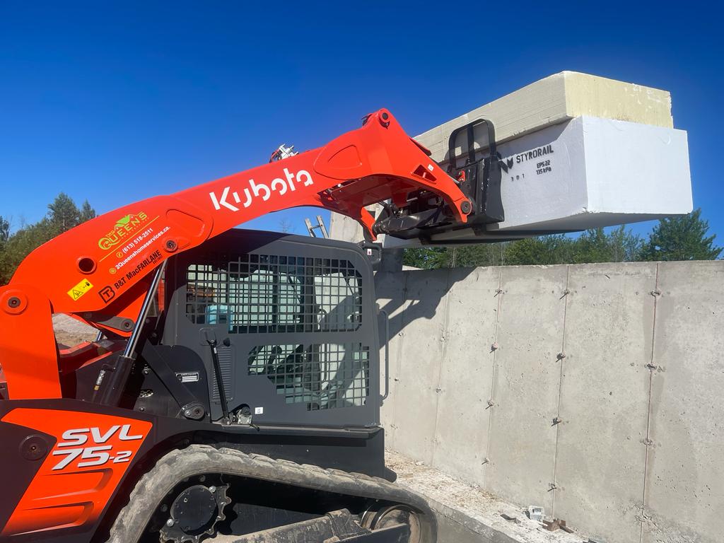 Close-up image of a Kubota SVL75-2 compact track loader in operation, lifting a large white Styrofoam block against a clear blue sky. The loader, marked with the logo 'QUEENS', demonstrates its power and precision on a construction site with concrete walls in the background.