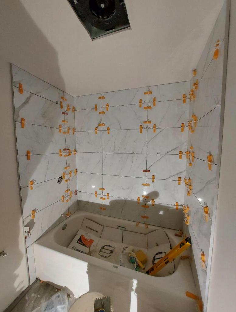 bathroom mid-renovation with large marble-patterned tiles being installed on the walls around a bathtub. The tiles are held in place with yellow leveling spacers, ensuring even alignment and spacing, a testament to the meticulous attention to detail in modern bathroom design