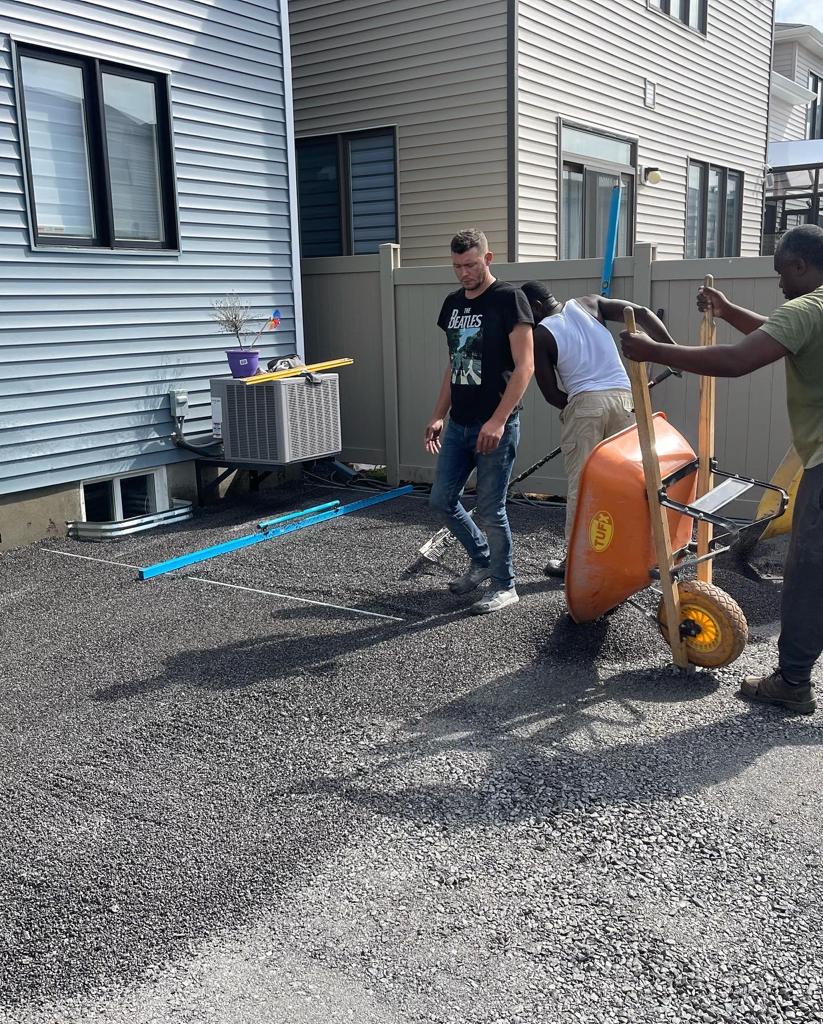 Landscaping experts actively preparing for an interlocking pavement project in Ottawa, with one person transporting gravel in a wheelbarrow and another smoothing it out, in the setting of a suburban home's outdoor space