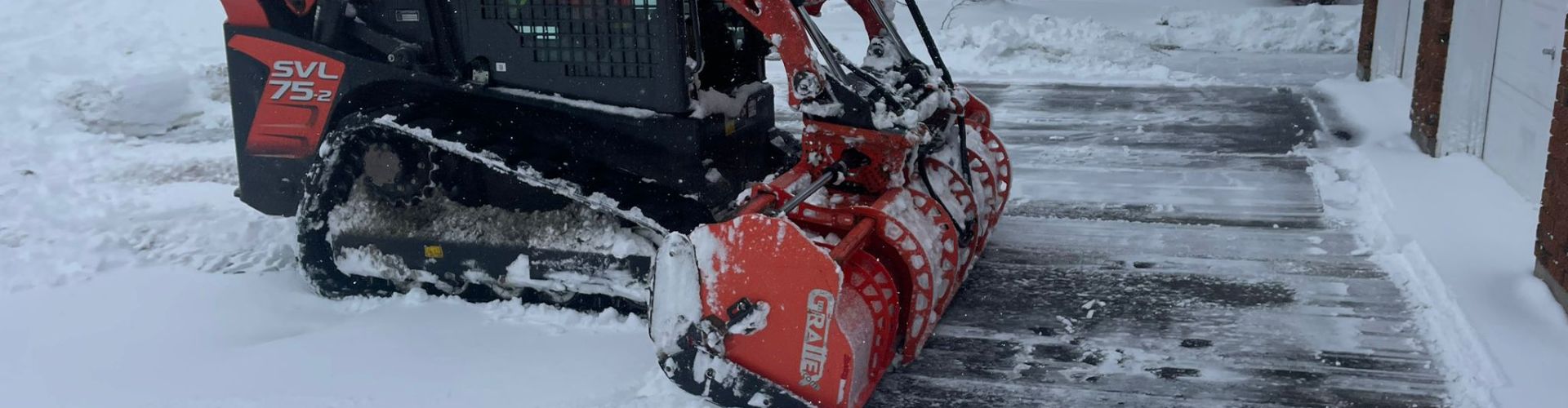 In the midst of a snowfall, a compact Kubota track loader equipped with a red snow bucket clears a residential driveway, showcasing effective snow removal during winter weather conditions.