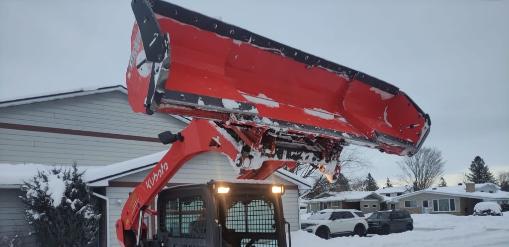 A Kubota snow plow attachment raised high, covered in snow against the backdrop of a residential neighborhood in Ottawa, indicating active snow removal services.