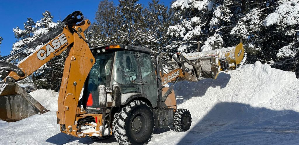 A sturdy CASE backhoe loader is actively removing snow in a wooded area. The scene showcases the machine's robust construction and the efficiency of snow management in a wintry landscape