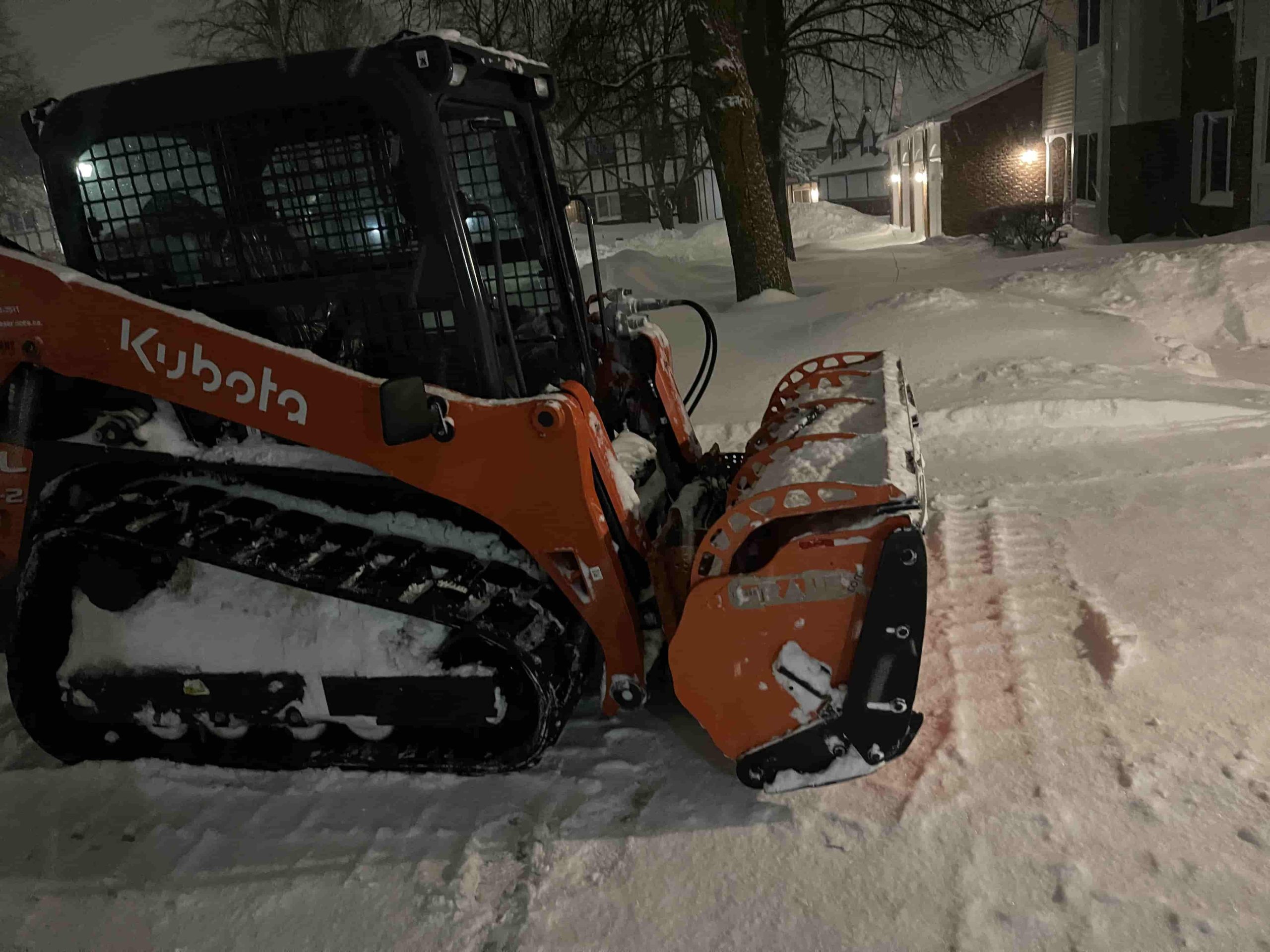 A vibrant orange Kubota skid steer loader is actively removing snow in a commercial area.