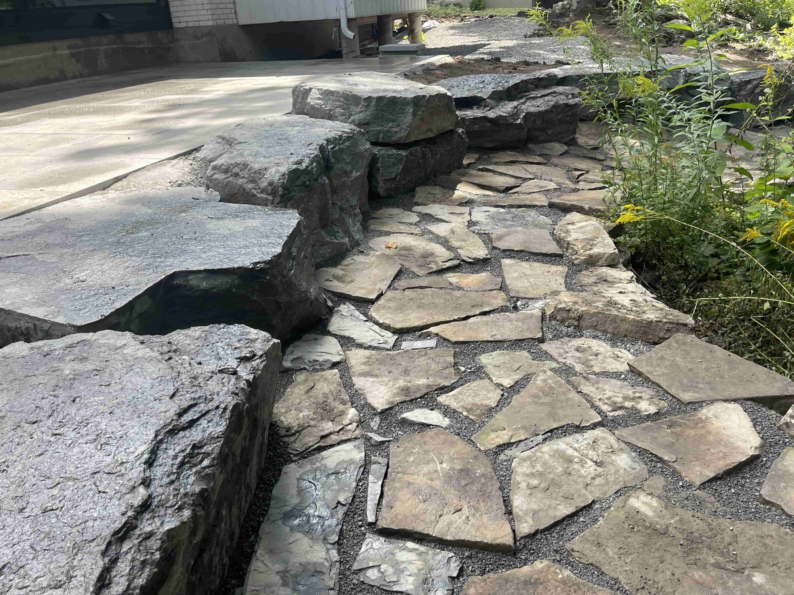 A skillfully installed flagstone pathway winds through a verdant garden, bordered by large boulders and lush foliage, showcasing the quality craftsmanship of professional flagstone services.
