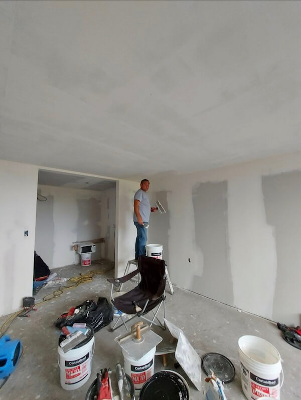 A worker stands on a platform with a trowel in hand, presumably applying joint compound to the seams of drywall—a process known as mudding. The walls are partially finished with visible patches of compound. Various tools and materials, such as buckets of compound, a utility knife, and a mixing paddle, are scattered around the room. The environment is typical of an in-progress construction site with unfinished details and an array of equipment. The worker appears focused on the task, indicative of a professional service at work.