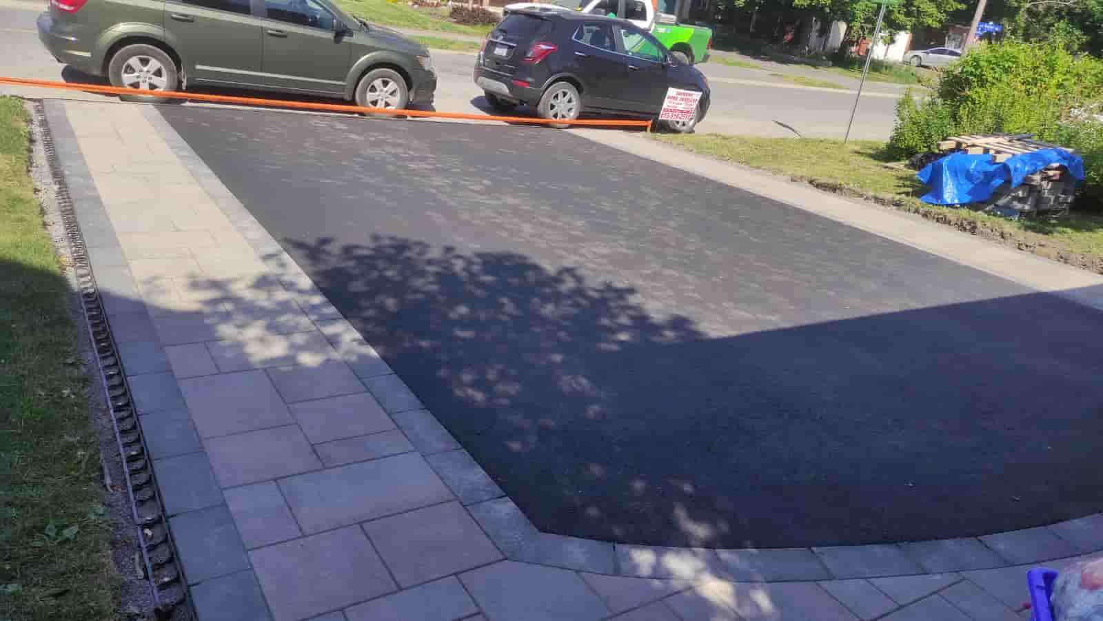 A new asphalt driveway installation with a neatly placed interlocking tile sidewalk. The contrasting colors of the grey tiles and dark asphalt create a clean and modern look.