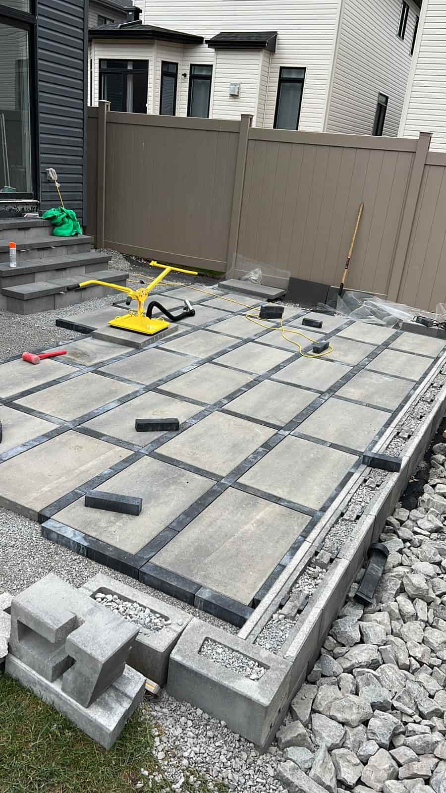 Installation of interlocking tiles in shades of gray on a residential patio, with construction tools, edging units, and a compacting machine on site.